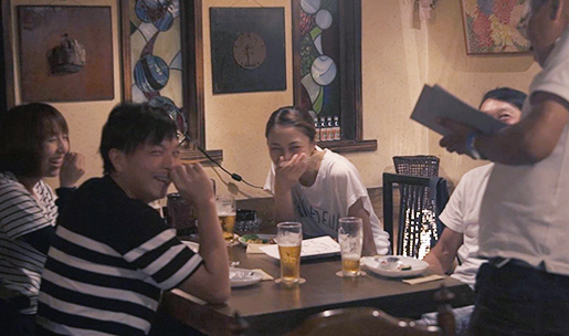 生ビールが置かれたテーブルで4名の人たちが店員の人と談笑している写真