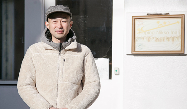 ゲストハウス「Nikko Inali sleping」の入り口前に立っている天野琢也さんの上半身を写した写真