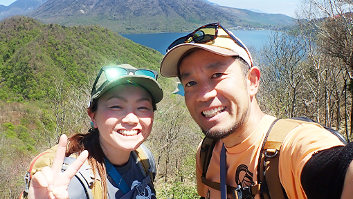 奥に山々と湖が見える社山で旦那さんと記念撮影をしている室町悦江さんの写真