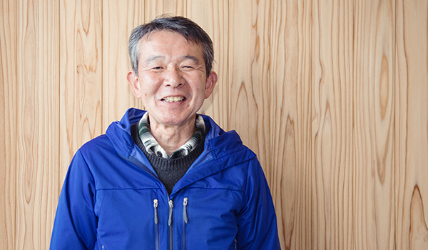 青いジャンバーに黒いセーター、シャツを着た山田徹さんが笑顔で立っている様子を正面から写した写真