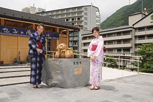 白地にラベンダー色の浴衣をきた女性と紺地に赤色の帯を巻いた女性が「鬼怒子の湯」と書かれた暖簾が掛けられている施設を背景に鬼の銅像の前で記念撮影をしている写真