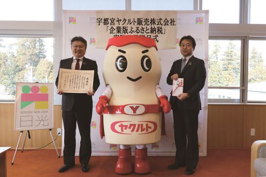 日光市のバックボードの前で、感謝状を持った宇都宮ヤクルト販売株式会社の男性とヤクルトマン、市長が一緒に記念撮影をしている写真