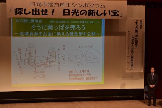 「探し出せ!日光の新しい宝」と書かれた幕の下に講演の内容がスクリーンに映し出され、右側に男性が立っている写真