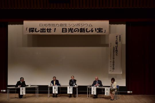 「探し出せ!日光の新しい宝」と書かれた幕の下に机と椅子が設置され、4人の男性と1人の女性が席に座っている写真
