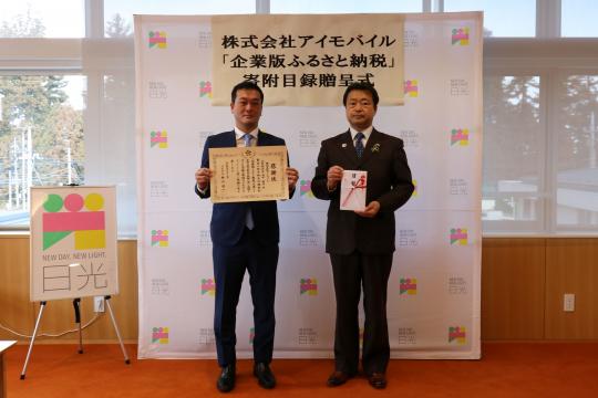 日光市のバックボードの前で、感謝状を持った株式会社アイモバイルの男性と市長が一緒に記念撮影をしている写真
