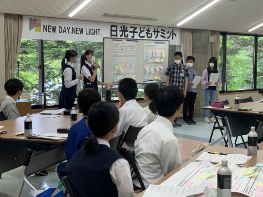 「日光子供サミット」と書かれたボードの前で、5人の学生が資料が貼られたホワイトボードの横に立ち意見交換をしている写真