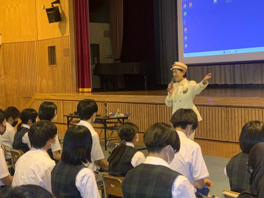大沢中学校体育館の舞台前で、制服を着た田沼下今市駅長が生徒の前で講話をしている写真