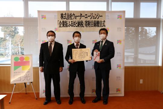 日光市のバックボードの前で、感謝状を持った株式会社ウォーターエージェンシーの男性2名と市長が一緒に記念撮影をしている写真