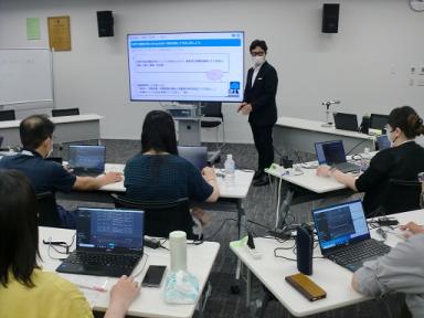 前方に設置されたモニターの横に男性が立って話をしており、席に座りノートパソコンを机に置いて話を聞いている参加者の方々を後ろから撮影した写真
