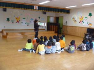 絵本を持ち立って読み聞かせをしている先生と、床に座って聞いている子ども達の写真