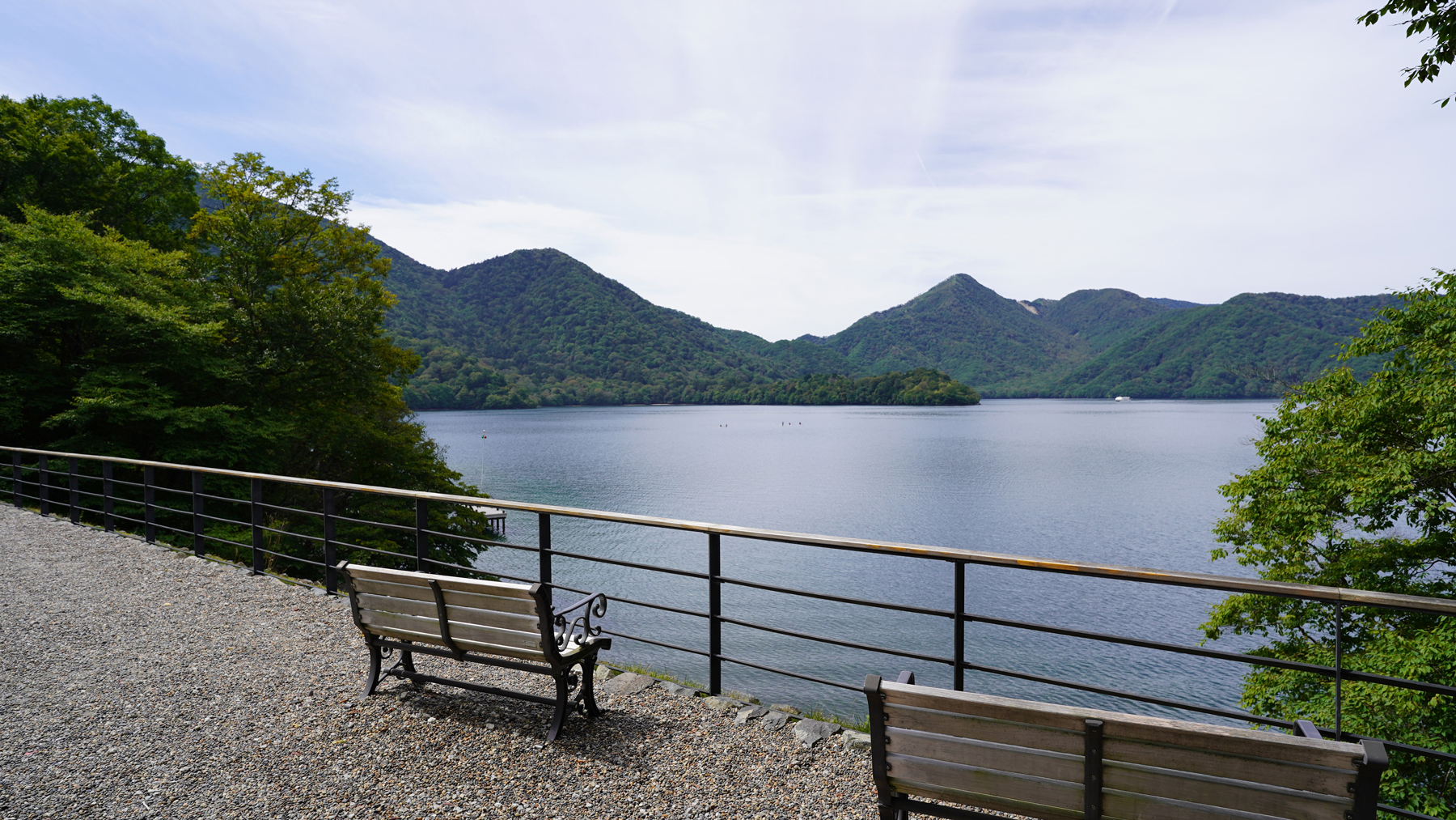 湖のほとりに設置されたベンチの後ろから見える湖と山々の写真