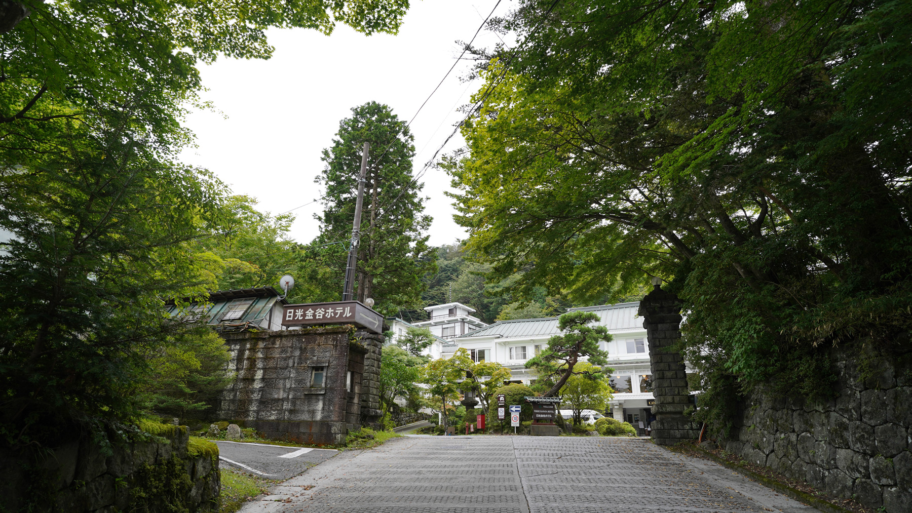 緩やかな上り坂右側の石造りの壁が続く先に門があり、左側に「日光金谷ホテル」の看板、奥には白い外壁のホテルが見える写真
