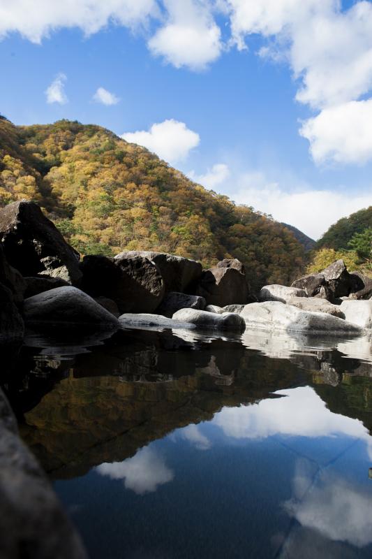 黄色や赤に染まってきている山々を背景に、晴天の青空が水面に反映している岩に囲まれた露天風呂の写真