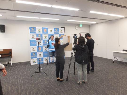 イベントのパネルが設置され、3人の撮影関係者の前で水色のTシャツを着た市長が国体冬季大会100日前イベント動画撮影を行っている様子の写真