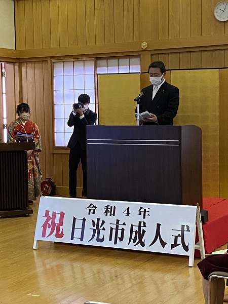 「令和4年祝日光市成人式」と書かれた看板が演台前に設置され、金屏風の前で挨拶をしている市長の写真