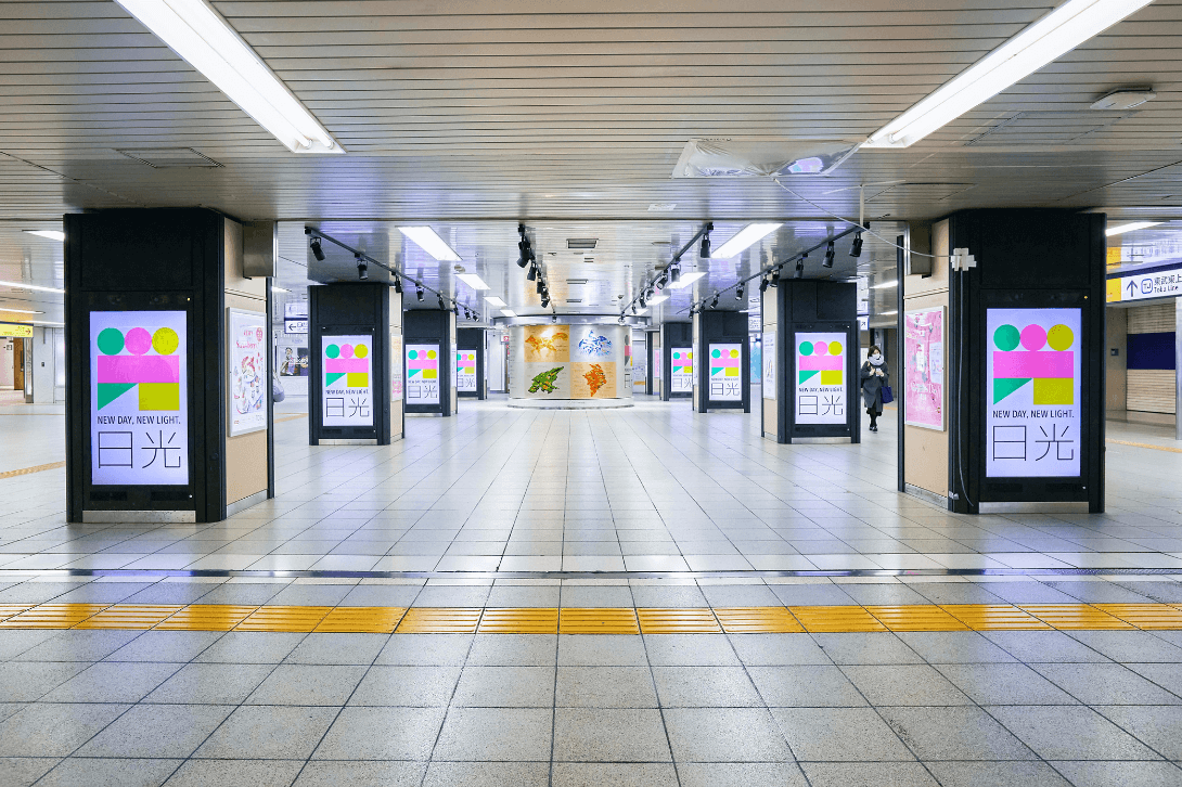 駅ホーム内の柱にNEW DAY, NEW LIGHT. 日光シンボルマークが映し出されているデジタルサイネージを写した写真