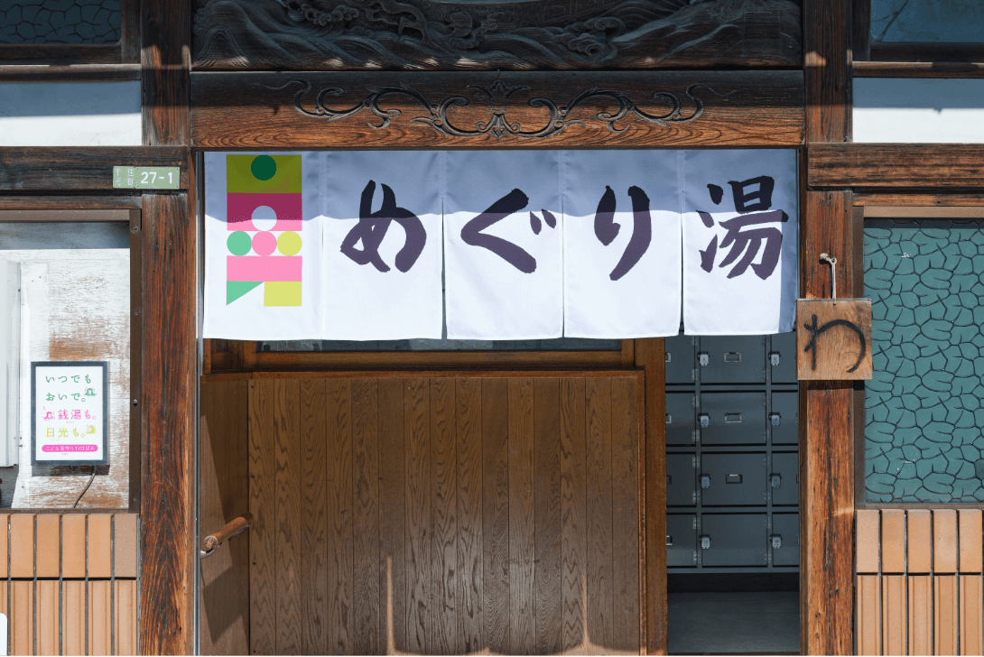 NEW DAY, NEW LIGHT. 日光シンボルマークと「めぐり湯」と書かれた暖簾が入り口に掛かっている写真