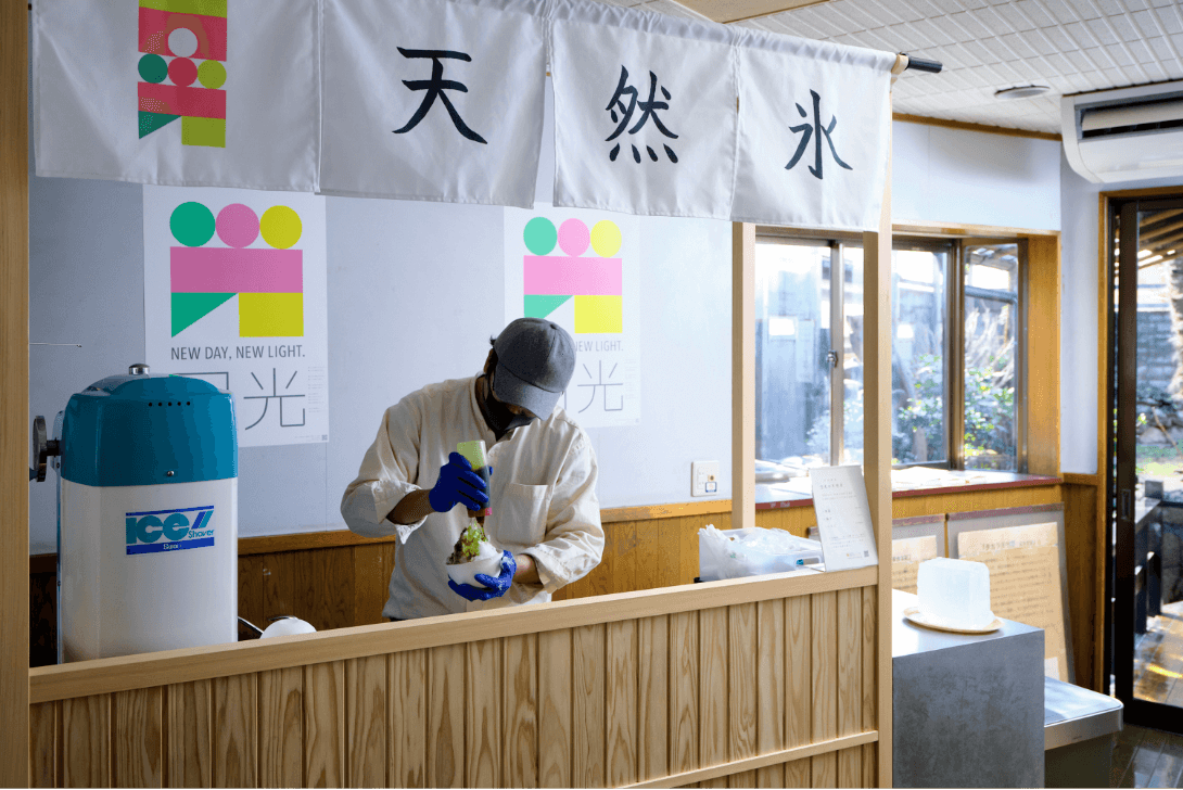 後ろに「NEW DAY, NEW LIGHT. 日光」のポスターが貼られ、「天然水」と書かれた暖簾の下で天然氷を使いかき氷を作っている男性の写真