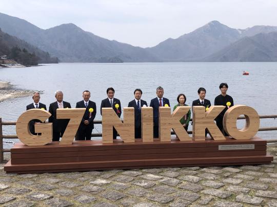 山と池をバックにG7モニュメントの後ろに市長と関係者8人が並んでいる写真