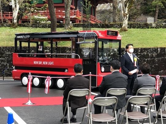 パイプ椅子に参加者たちが座り、赤と黒のデザインで小型のバスのような形をしたグリーンスローモビリティの前に市長が立って挨拶をしている出発式の様子写真