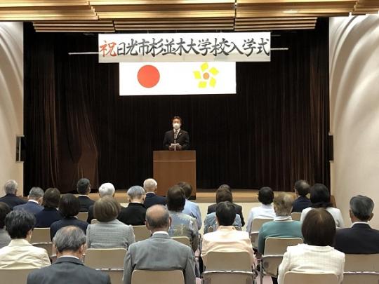 演壇で挨拶をしている市長の話を聞いている参加者達を後方から写した杉並木大学校入学式の様子写真