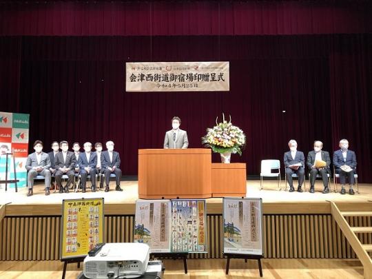 舞台下に3枚のパネルが置いてあり、演壇で話をする市長と両端に関係者が座っている会津西街道御宿場印贈呈式の写真