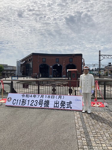 白い制服を着た市長が「c11形123号機出発式」のパネルの横に立っている出版式の様子写真