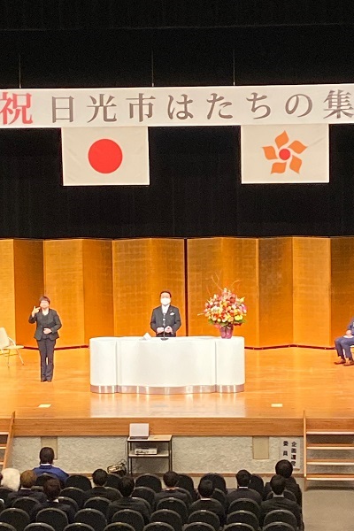 「はたちの集い」で演壇の金屏風の前で挨拶する市長の言葉を聞いている参加者の人たちの写真
