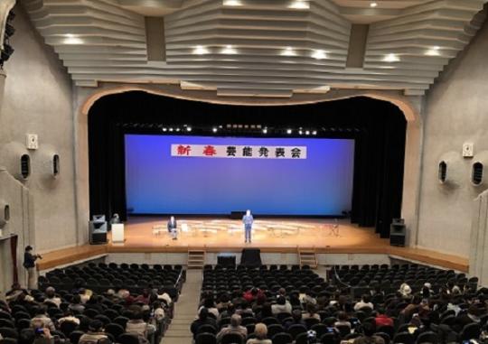 「新春芸能発表会」と書かれ大勢の参加者が集まっている新春芸能発表会の様子を2階席から舞台方向を写した写真