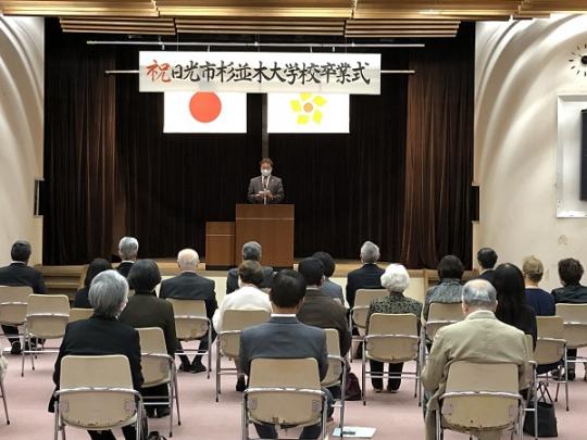 参加者が椅子に座り、演壇で市長が挨拶をしている日光市杉並木大学校卒業式の様子を後方から撮影した写真