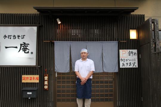 そば屋の店頭ののれんの前に立つ頭にバンダナを巻いた男性の写真