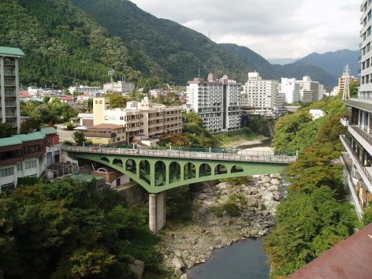 山々と複数の建物に囲まれている緑色の橋とその下を川が流れている様子の写真