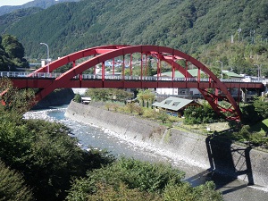 緑の山々を背景にした赤色の大橋とその下を穏やかに流れる川の写真