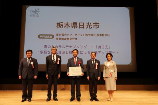 ステージにかけられたスクリーンの前に、認定証授与式にて中央の男性が賞状を持ち、両脇にそれぞれ2名ずつ関係者が並び記念撮影をしている写真