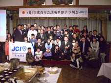 宴会場の舞台前で、顔が見えるように4列ほどに並び老若男女が記念撮影をしている写真