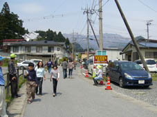 住宅街の道路を行き交う人と作業服などを着た関係者がその様子を見守っている写真