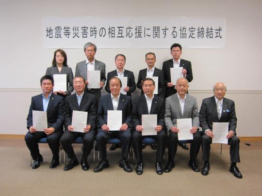 「地震等災害時の相互応援に関する協定締結式」と書かれた横断幕の前でスーツを着用した男女が2列に並び協定書を両手で持ち記念撮影をしている写真