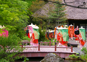 華やかな衣装の侍女達が薄い緑色の布をつけた笠に手をあて橋の上を渡っている平家大祭の写真