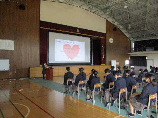 体育館の舞台前に設置されたスクリーン画面の横で話をしている講師と椅子に座って話を聞いている大勢の生徒たちの写真