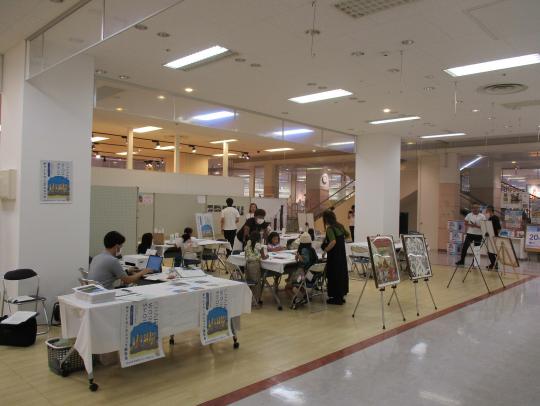 掲示板やイーゼルスタンドに立てかけられた看板や絵に囲まれた商業施設内の一角に、長テーブルと椅子がいくつも置かれ、参加者や関係者を撮影をした写真