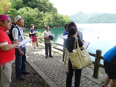 参加者の方々が湖の畔で資料を手に持ちながら見学している写真