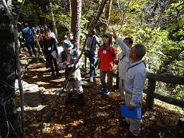 山の中で男性が手を挙げジェスチャーをしながら説明し、参加者の方々が上の方に注目して話を聞いている写真