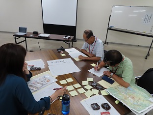 長机が設置された会議室で、資料や地図を机に並べ参加者の方がメモを取っている写真