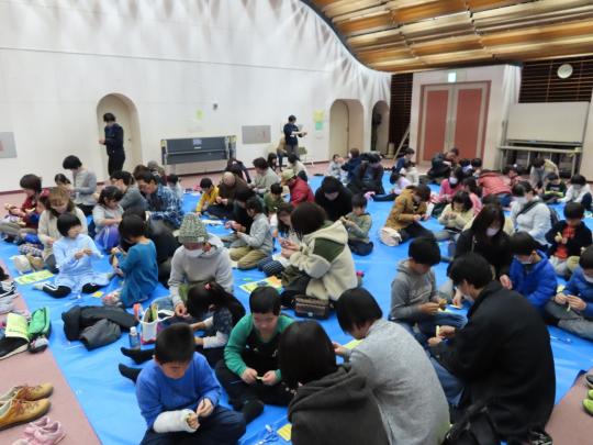 大きなホ-ルでブルーシートが敷かれた場所に座って体験教室に参加している人々の写真