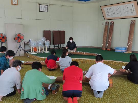 1段上がりのある室内で講師の方が箏の手本を見せ、箏を置き床に座って弾き方をまねている子ども達の写真