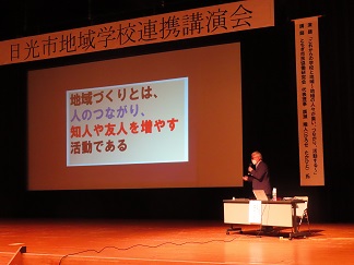 壇上で男性がマイクを持っていて、スクリーンに「地域づくりとは、人のつながり、知人や友人を増やす活動である」という文字が映し出されている写真