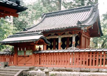 石が積み上げられた土台の上に朱色で屋根が付いた縦格子の柵の奥に建っている、全体が朱色で切妻屋根の二荒山神社別宮本宮神社本殿の写真