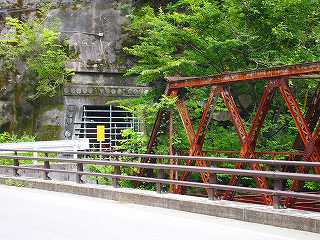 道路横に赤いトラス橋、その奥に入り口が鉄の棒で封鎖されいている小滝坑跡の写真