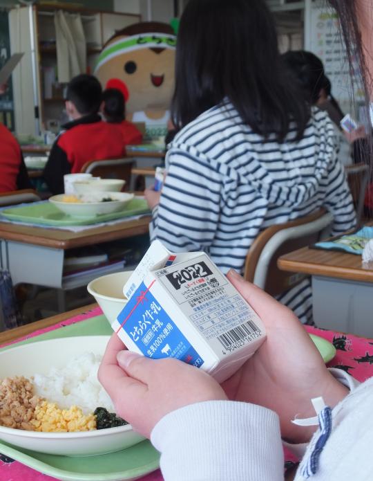 配膳された給食から牛乳パックを両手で持って取り上げている写真