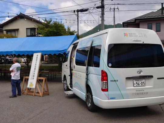足尾納涼祭の広場に青いテントが張られ、移動期日前投票所体験会と書かれた看板が真ん中にあり、その横に白いマルチタスク車両の扉が開いてる写真
